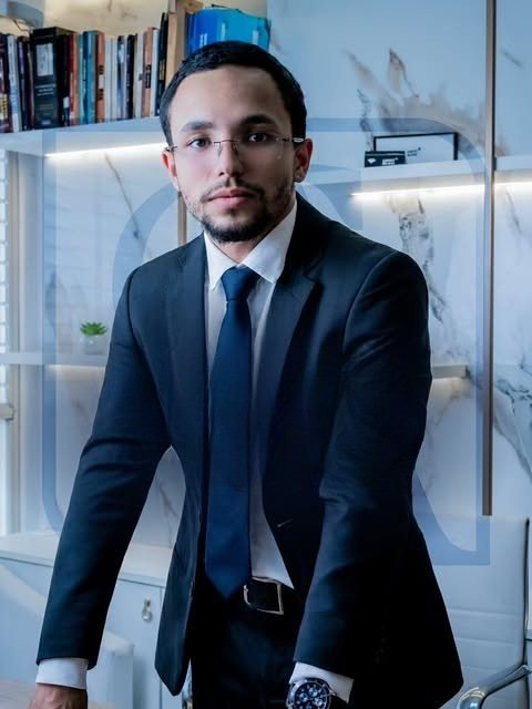 Allan Soares em seu escritório, apoiado na mesa, com estante de livros ao fundo
